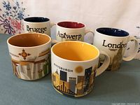 Five Starbucks mugs arranged to show exteriors with city names and designs portraying London, Brugge, Antwerp, Phoenix, and Pittsburgh