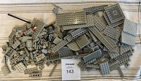 Top view of a pile of assorted grey LEGO plates and parts on a light striped fabric background, with a MaxSold lot card visible