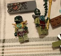 Close-up of two green Croc Tribe LEGO minifigures with armor and accessories including spear and detailed torso printing.