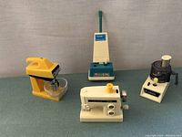 Five miniature household appliance models: blender, food processor, vacuum, sewing machine; shown on flat surface with neutral background