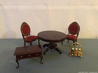 Front view of two miniature chairs with red velvet upholstery, round wood pedestal table, small side table with drawers, and brass drinks rack with miniature bottles.