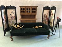 Formal miniature living room scene showing the dark green upholstered couch with two brass instruments, four Asian decorative panels behind, wood bookcase in background, and metal camera on tripod.
