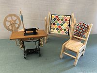 Miniature wooden sewing machine with a black sewing machine, metal legs, and spinning wheel, quilt display with needlepoint quilt, and wooden chair with needlepoint seat and back.