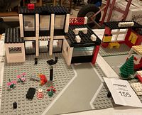 Front view of LEGO police station building, small accessories including flowers and traffic cone, and partial view of bus stop area.