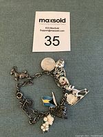 Photo of sterling silver charm bracelet with seven distinct charms against a teal fabric background with a MaxSold lot number card above.