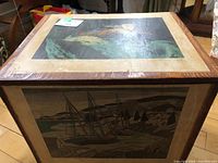 Wooden cube trunk showing decorative illustrated panel of a woman on the top surface and part of a scenic panel on the side.