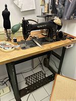 Black antique Singer sewing machine mounted on metal treadle stand with wooden top, showing machine and sewing accessories.
