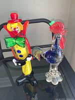 Both clown and rooster glass figurines side by side on glass table.