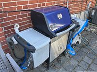 Overall view of the Weber BBQ grill showing left side shelf covered with a grey fabric cover and propane hookup connection on the back wall.