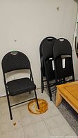 Four black folding chairs, one open and three stacked, with metal frames and dark upholstered seats and backs. Located indoors on tiled and carpeted floor.