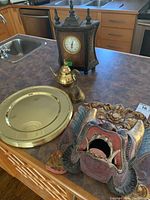 Wood mantel clock, brass bell and teapot, decorative carved wood mask with open mouth and floral detail, plastic gold charger plates stacked