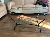Oval metal frame coffee table with clear glass top and shelf, shown on hardwood floor with beige couch and chair in background