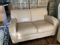 Front view of beige leather loveseat couch showing two cushions and rounded arms against background of packed boxes.