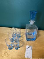 Photo showing the decanter alongside six small liqueur glasses arranged on a wooden surface with a teal background.