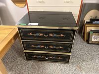 Front view of the three drawer black dresser showing painted floral designs on drawer fronts and gold trim.
