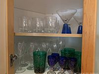 Two shelves showing an assortment of glasses including clear whiskey glasses, stemmed wine glasses, green decorative glasses, smaller blue decorative glasses, and blue-stemmed martini glasses.