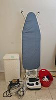 Gray ironing board with metal frame next to white hamper and three irons on floor in front, and red laundry basket.