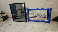Two wall mirrors leaning against a beige cinder block wall on tiled floor. The left mirror has a simple black metal frame and the right mirror has a blue plastic frame with zigzag edges.