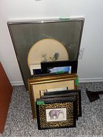 Collection of nine framed prints plus one framed fiber art piece of varying frame styles and sizes leaning against a wall.