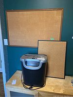 Fellowes OS-8Cs paper shredder on wooden cabinet with two cork bulletin boards behind it on blue wall.