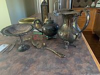Photo showing all five items together on countertop: large jug, teapot, tongs, pedestal bowl, and serving utensil.