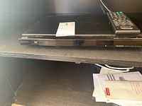 Sony black DVD player placed on a shelf with remote control on top, close-up view showing front panel with Sony and DVD logos.