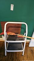 Two-step ladder with broom and dustpan on top, showing details of ladder steps and broom bristles.