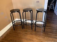 Three metal stools with round seats and tubular legs positioned against a wall on hardwood floor.
