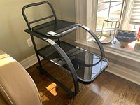 Side table with black metal frame and two black glass shelves, placed near a window with natural light highlighting the scratches on the glass surface.