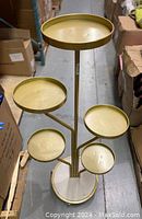 Front view showing five circular trays of varying sizes and heights mounted on a gold central pole, standing in a warehouse environment.