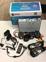 Full set of Anni security system components, including box, DVR, 4 cameras, remote, power adapters and cables laid out on a floor.