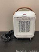 Front view of white De'Longhi Capsule ceramic heater showing grille and brown handle. Black power cord visible coiled beside unit.