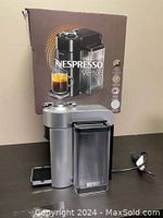 Side view of the Nespresso Vertuo coffee machine in silver with water tank and drip tray, standing in front of original box.