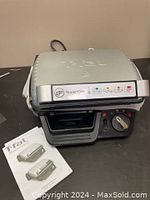 Front view of the closed T-FAL Super Grill showing control dial, power cord, and brand logo on lid with user manual beside it.