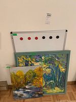 Photo of two original paintings on board leaning against wall with a magnetic whiteboard and magnets above them.