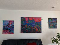 View of three original abstract paintings on wall above black leather couch, colors include pink, purple, green and orange in organic shapes.