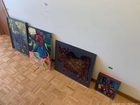 Four paintings arranged on floor against wall showing size and framing: floral still life framed on board, cow painting on canvas, abstract canvas painting, and small unframed abstract painting.