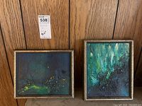 Pair of framed abstract paintings on canvas board with wood panel background. One is horizontal with darker blues, greens, and yellow accents. The other is vertical with greens and textured surface.