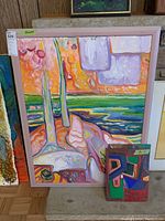 Full view of the framed large painting on board with a colorful abstract landscape in an orange and purple color scheme, pink frame, with two smaller unframed paintings leaning against it.