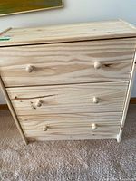 Front view of an unfinished pine dresser with three drawers and round wooden knobs sitting on carpet.