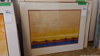 Framed watercolor painting with a landscape scene showing a blue water body, orange-brown land with trees, and a yellow sky.