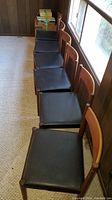 Six teak frame chairs with black vinyl seats arranged in a row on carpeting