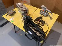 Wide view of all tools on yellow tables including Black & Decker sanders, jig saw, planner, Stanley stapler, and Dremel with case and attachments.