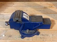 Side view of the blue metal vise sitting on a wooden surface, showing the jaw and handle clearly.