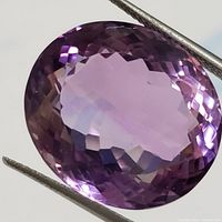 Close-up photo of an oval faceted amethyst gemstone held by tweezers, showing deep purple color and multiple facets.
