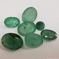 Seven small to medium natural emerald gemstones with varied oval and rounded cuts, showing natural inclusions and typical green color, placed on white background.