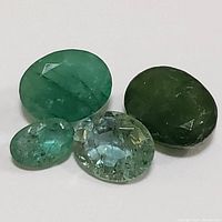 Four oval faceted emerald gemstones of varying green shades arranged on a white surface.