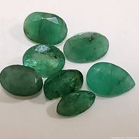 Seven loose genuine emerald gemstones varying in shape and size, displayed on white background showing natural inclusions and color variations.