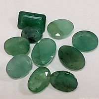 Photo of 11 genuine emerald gemstones in various oval cuts and one rectangular cut showing color, shape, and typical inclusions.