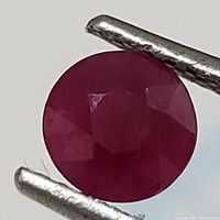 Close-up photo of the round cut genuine ruby gemstone held by tweezers showing facets and rich red color with some inclusions.